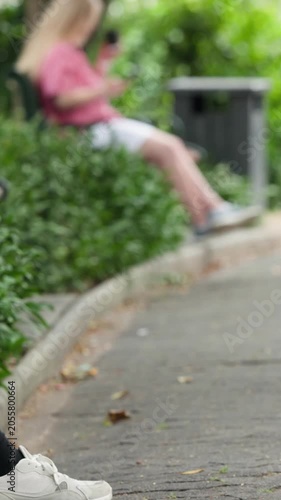 Vertical video: Scrolling blonde teen in pink top using phone on path while bicycle enters, passing