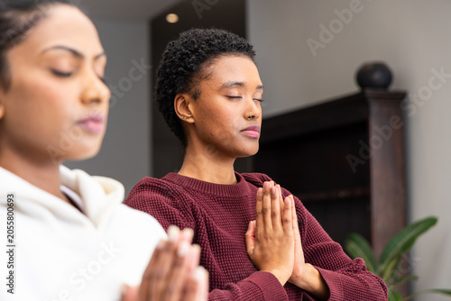 Indian, African American women meditating with hands in prayer at home, maroon sweater white hoodie