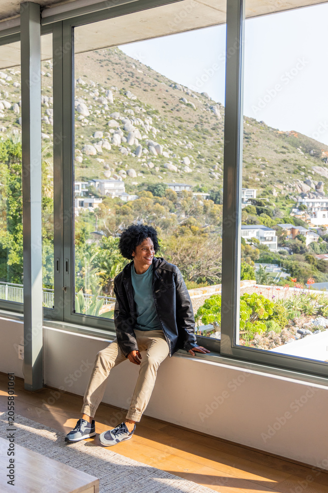 Naklejka premium African American man in 20s sitting on wide windowsill by glass walls, sunlight on jacket