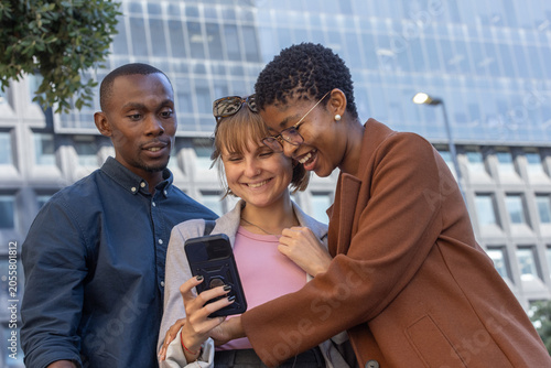 Diverse friends leaning in urban plaza, sharing smartphone with dark case, wearing blazer and coat