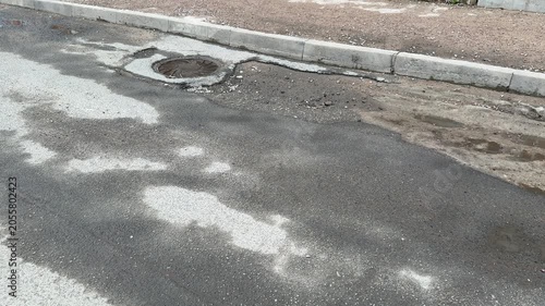 Worn asphalt road with potholes showing curbside emergency lane, gravel shoulder, standing puddles, cracked surface, localized