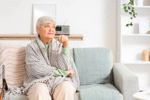 Beautiful thoughtful senior woman with plaid and eyeglasses sitting on sofa a...