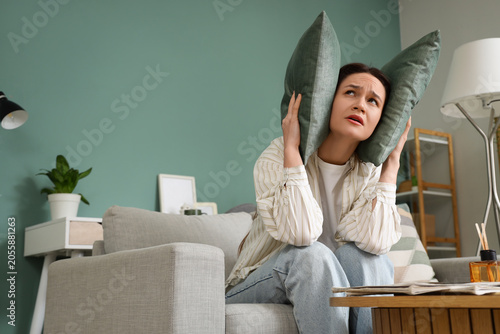 Fotografi Young woman covering her ears with pillows because of noisy neighbours on sofa a