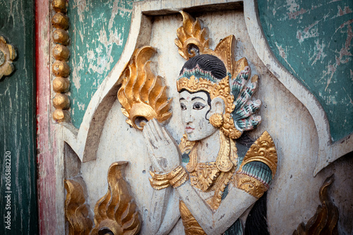 Balinese wood carving of a serene female deity with hands in prayer, wearing ornate ancient costume and gold leaf details on a weathered temple door in Bali, Indonesia