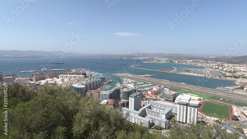 Wide angle shot from Rock of Gibraltar shows city skyline, marina, airport runway, blue bay, distant hills, bright daylight, clear sky, and lush foreground vegetation in sharp focus.