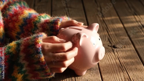 Person in colorful knitted sweater holding pink piggy bank on wooden surface in warm sunlight