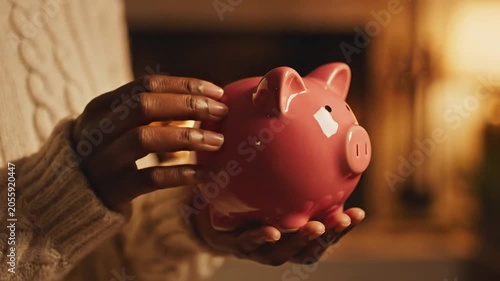 Person in cozy white sweater holding pink piggy bank in front of warm glowing fireplace