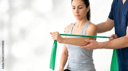 Professional physical therapist assisting a young woman with arm rehabilitation exercises using a green resistance band in a bright clinic setting.