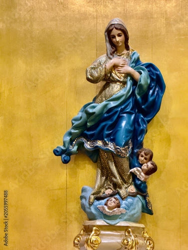 Religious statue of the Virgin Mary standing on clouds with angels, dressed in blue and white robes, against a golden background. Catholic devotional imagery symbolizing purity, grace, faith, 