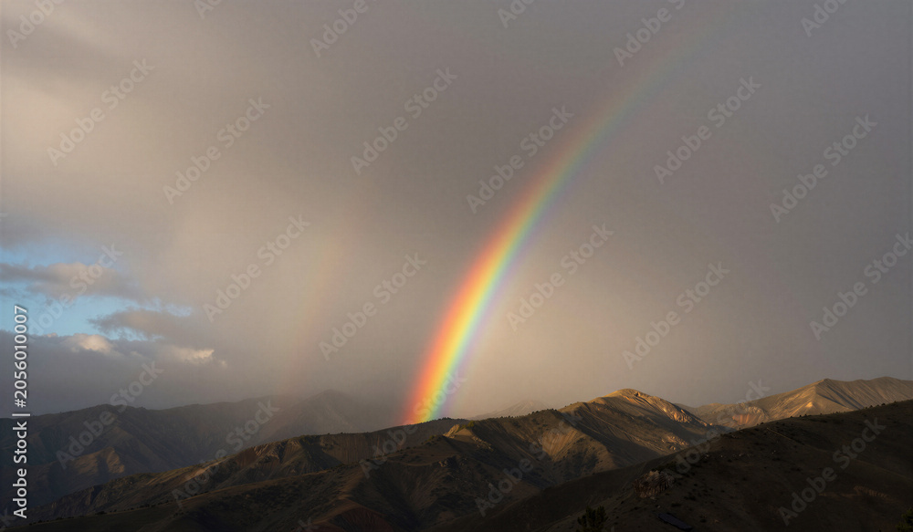 Naklejka premium Rainbow arches gracefully over rugged mountains. Clouds swirl behind, softening the sky's edge. Distant peaks stand bold beneath the natural display. Nature's calm beauty after stormy weather
