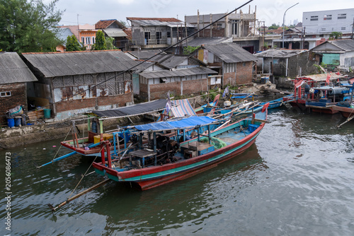 A high-angle view of colorful traditional wooden fishing boats docked along a waterfront in Cilacap, Indonesia, with a densely packed coastal settlement in the background.
