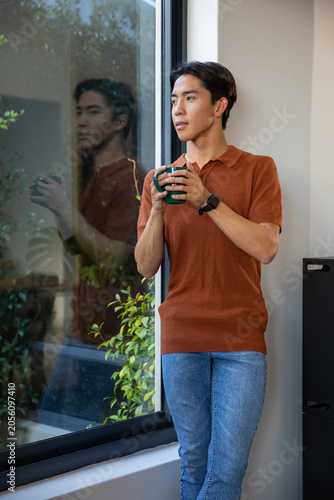 Adult Asian man wearing rust polo, jeans standing beside glass window holding green mug, copy space