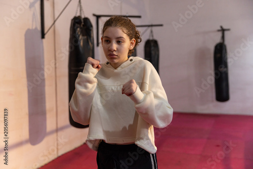 Teenage boxer practicing on red mats at gym in cropped hoodie and pants with hanging bags