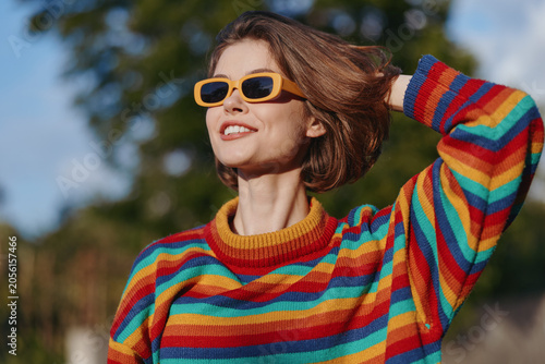 Fotografía Woman, smiling, rainbow sweater, yellow sunglasses, short hair, outdoor, sunny, casual: Young 20s female model in colorful striped sweater, happy and confident, looking up with hand in hair in park