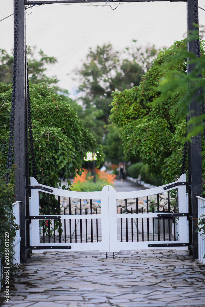 Fototapeta premium Garden gate framed by lush greenery opens toward peaceful pathway, gate creating elegant entrance in natural setting. Gate scene highlights symmetry, outdoor architecture, decorative detail. vertical