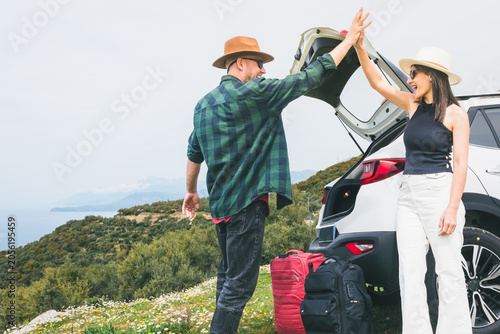 Fotografie Happy Travelers tourist couple hi5 stand put luggage to white rented SUV vehicle trunk on scenic Mediterranean viewpoint