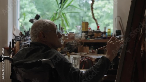 Senior artist on a wheelchair paints an oil painting on canvas in a studio