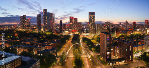 Aerial panorama over Manchester at twilight, featuring illuminated skyscraper towers, light trails on the Princess road below
