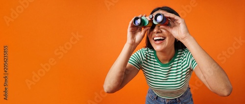 Woman looking through binoculars smiling excited on bright orange background, happy young female searching for future opportunities, creative discovery concept, curious explorer seeking clear visions.