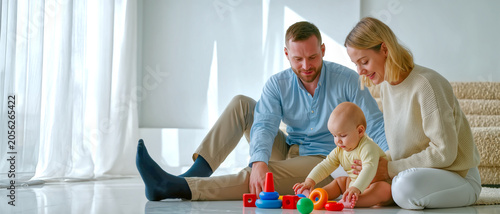 Young parents sitting on a sunlit floor, watching their infant baby play with colorful stacking toys in a bright, peaceful home environment, capturing a warm and loving family moment.