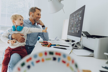 Playful son with wireless headphones sitting on lap of his father working at home office
