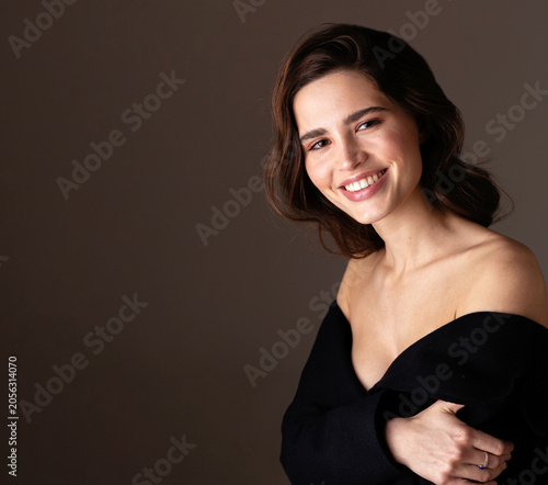 A smiling young woman with long wavy brunette hair poses in an elegant off-shoulder black dress against a warm neutral studio background. Soft lighting enhances her radiant skin and natural beauty