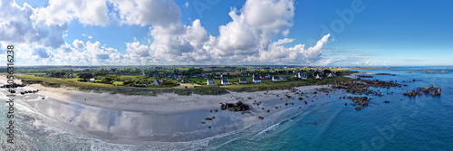 Ein weites Panorama vom Plage de Boutrouilles mit hellem Sand, türkisblauem Meer, den typischen Granitfelsen und einer ländlichen Siedlung unter markantem Wolkenhimmel.