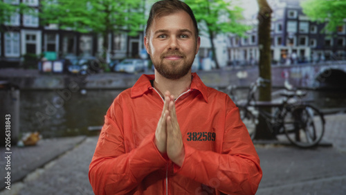 Foto Man in orange prisoner jumpsuit numbered 38258 with palms pressed together praying beside a canal on an amsterdam street near parked bicycles and a bridge; hopeful plea redemption