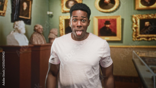 African american man grinning with hands raised, fingers splayed and tongue out while standing before framed portraits and classical busts in a gallery building; playful joy and curiosity.
