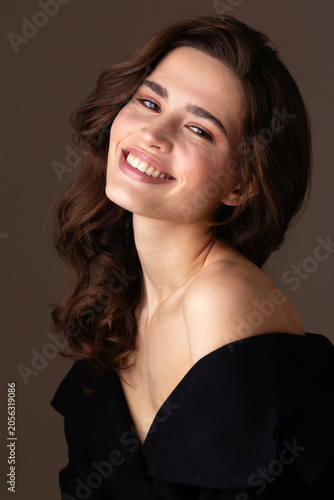A smiling young woman with wavy brunette hair poses in an elegant black off-shoulder dress against a soft neutral background. Her natural makeup, radiant expression create a sophisticated portrait.