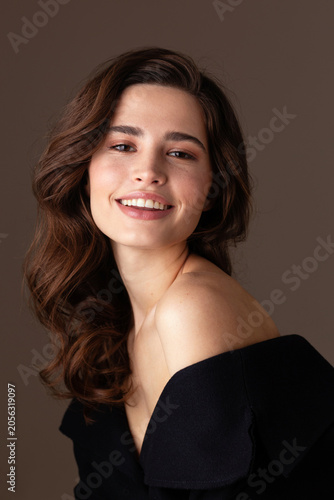 A beautiful young woman with wavy brunette hair smiles gently at the camera while posing in an elegant black off-shoulder dress. Soft natural makeup create a timeless beauty and fashion portrait.