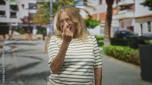 Schilderij op canvas Senior blonde woman winks and beckons with fingers, bare hand forward, wearing striped shirt in a sunlit pedestrian street with palm trees; playful teasing