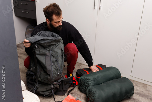 Fotografie Young bearded man packing large tourist backpack with sleeping bags and camping gear at home