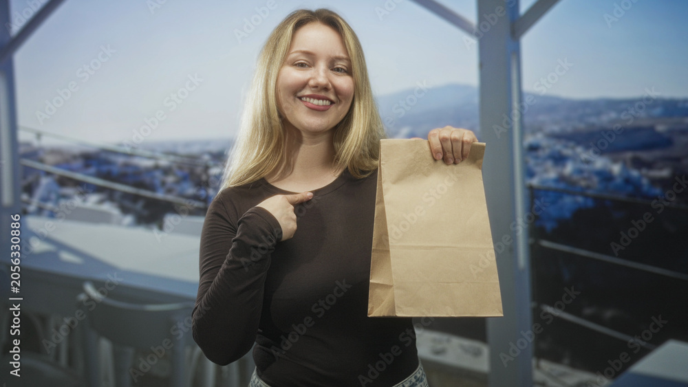 Fototapeta premium Smiling blonde woman pointing finger to chest while holding paper bag on building balcony overlooking town; warm pride.
