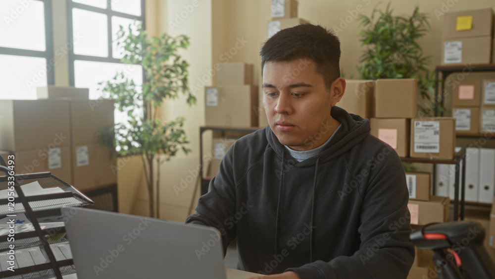 Fototapeta premium Man with laptop leaning back with hands behind head in office building surrounded by cardboard parcels and packing shelves; relief productivity.