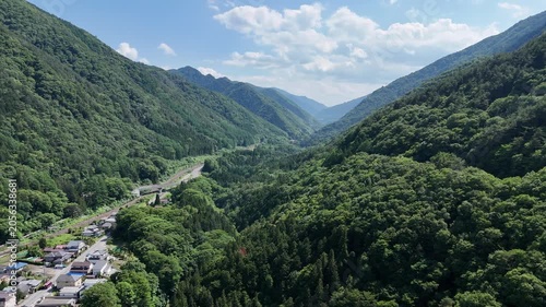 Lush green mountains frame a winding valley. River flows beside a small town. White clouds drift above the peaks. Vibrant foliage blankets every slope. Serene, untouched nature draws the viewer in