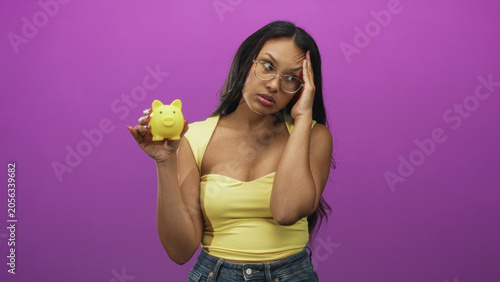 Принти на полотні Woman holds yellow piggybank, hand on temple, wearing glasses and yellow top in studio; saving dilemma concern