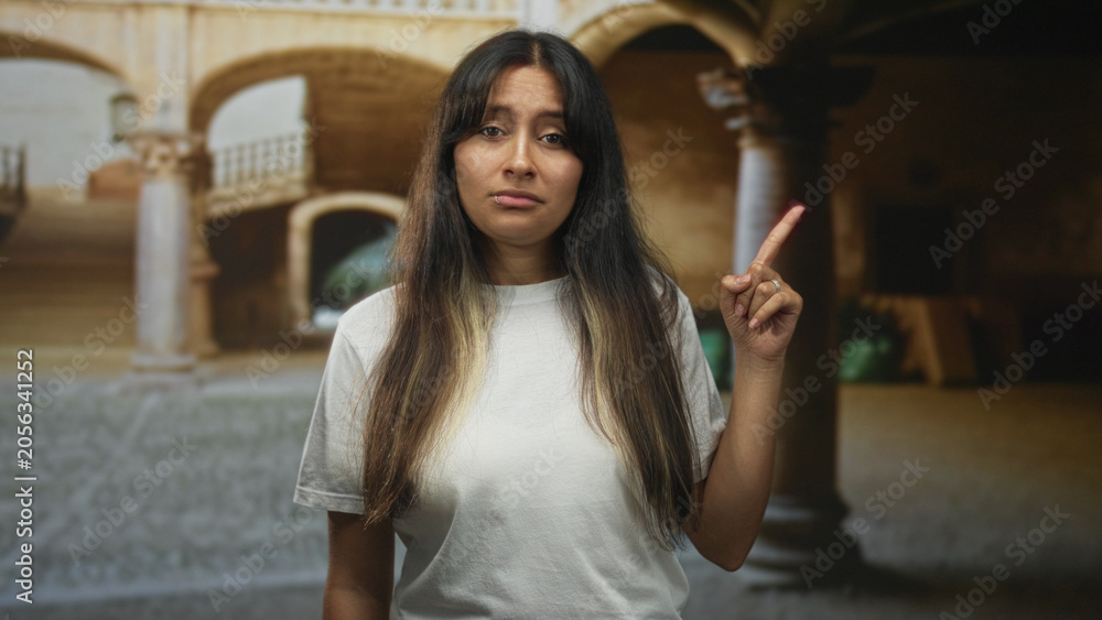 Fototapeta premium Woman points finger upward in old town building courtyard, white t shirt and lip piercing visible; skepticism caution.