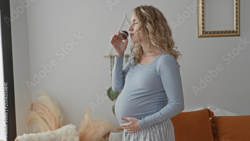 Canvastavla Young pregnant woman, blonde, holding a chocolate muffin near her lips with one hand and resting the other hand on her belly, slight smile in studio; maternal contentment