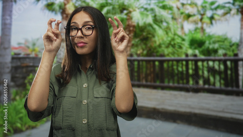 Fotografie Woman wearing glasses making air quotes gesture with both hands on a wooden boardwalk surrounded by palm trees in forest setting; playful skepticism