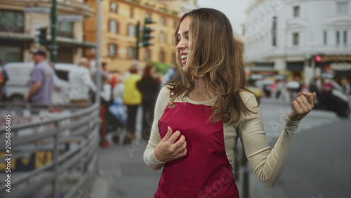 Tableau sur toile Hispanic woman in red apron gestures with hand, laughing and smiling while standing near a crosswalk on a busy city street; carefree joy