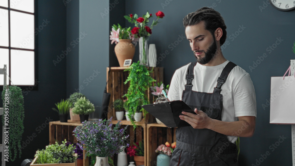Obraz premium Man florist in apron holding clipboard and writing with pen while pointing to flower arrangements in studio; serenity care.