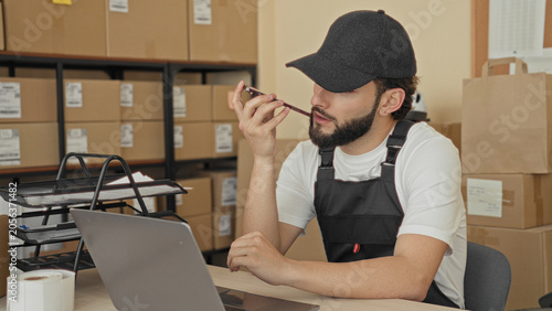 фотографія Man holds smartphone to mouth with hand while checking laptop and parcels on desk, surrounded by cardboard boxes on shelves in a building; focus productivity
