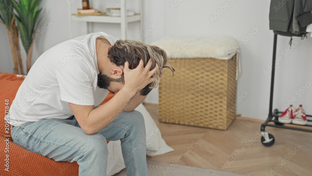 Fototapeta premium Man sitting on bed holding neck and head, clutching head with hands for headache in a building bedroom; anxiety stress fatigue.