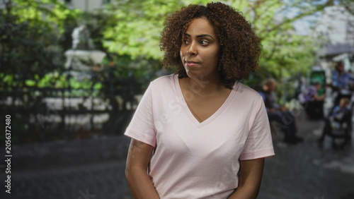 Fotografija Woman, young african american, with hands clasped at waist wearing pale pink v neck t shirt and curly hair, looking sideways while standing on city street beside park fountain; pensive reflection