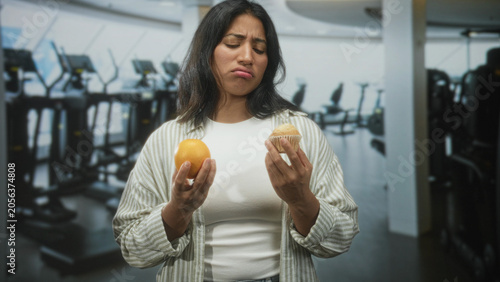 Schilderij op canvas Woman holds orange and muffin, weighing choice with hands and smiling then frowning inside a gym building; temptation choice