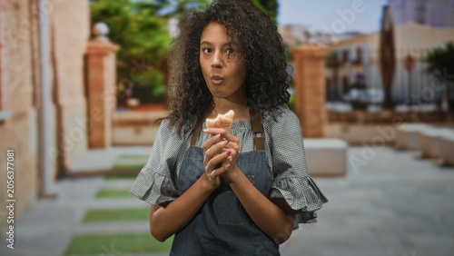 Photography Teenage woman cook in apron holding croissant, lips pursed while chewing on a street; quiet contemplation