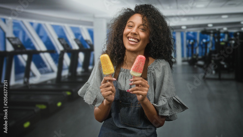 Tableau sur toile Woman with curly hair holding two colorful popsicles in her hands and smiling with eyes closed, apron visible inside a building gym with treadmills; carefree joy