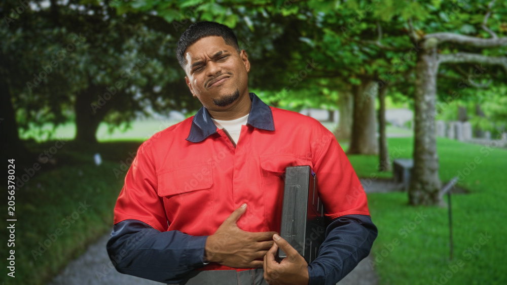 Fototapeta premium Hispanic man in red work uniform clutching chest while holding toolbox on a tree lined path in forest; pain duty resilience.