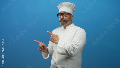 Fotografie Man chef in white uniform and hat pointing with hand in studio against vivid blue backdrop; confidence skill craft